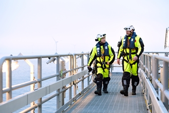 two men walking on a deck, dressed in high visibility clothing wearing helmets and a climbing harness