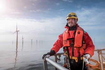 a man in protective clothing and harness is holding onto railing, posing for a picture with a row of wind turbines behind him