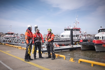 three men in protective clothing, wearing harness' are having a conversation in front of a bay full of boats