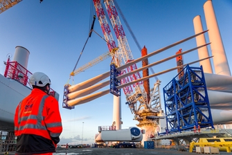 a person wearing a high visibility EDF jacket is overlooking a crane lifting three wind turbine blades