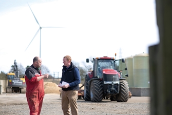 two men having a conversation on a farm, with a tractor close by and a wind turbine in the background